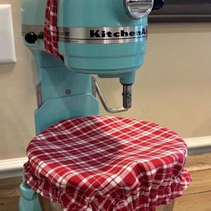 May include: A light blue KitchenAid stand mixer featuring a red and white plaid fabric cover on the mixing bowl and a matching red and white plaid fabric bow tied around the top of the mixer head. The mixer has a metallic silver bowl.