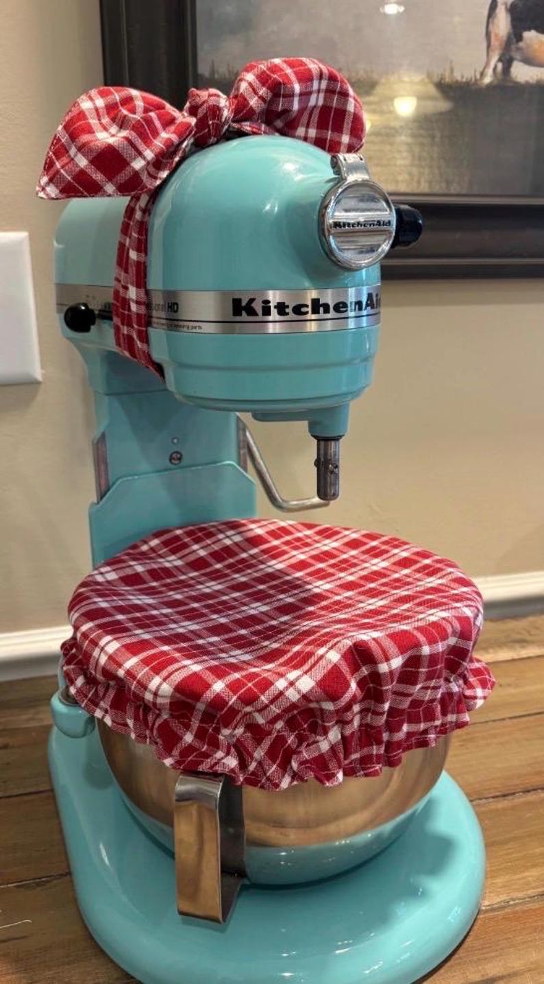 May include: A light blue KitchenAid stand mixer featuring a red and white plaid fabric cover on the mixing bowl and a matching red and white plaid fabric bow tied around the top of the mixer head. The mixer has a metallic silver bowl.