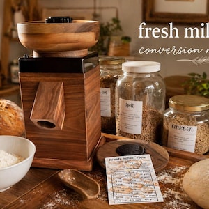 Puede incluir: Un molino de harina de madera con un cuenco de harina, una hogaza de pan y tarros de granos. La imagen incluye un imán de conversión con el texto "fresh milled flour conversion magnet". La escena está ambientada en una cocina rústica.