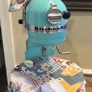 May include: A turquoise KitchenAid stand mixer featuring a fabric cover over the bowl and a matching fabric bow tied to the top. The fabric has a multi-colored patchwork pattern with floral, gingham, and striped designs in blue, yellow, and red.