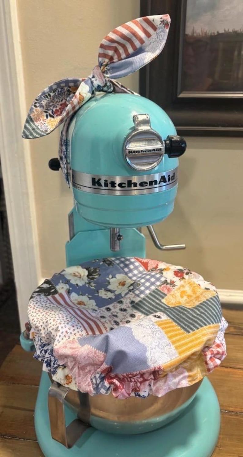 May include: A turquoise KitchenAid stand mixer featuring a fabric cover over the bowl and a matching fabric bow tied to the top. The fabric has a multi-colored patchwork pattern with floral, gingham, and striped designs in blue, yellow, and red.