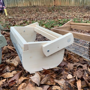 May include: A wooden bird feeder with a wire mesh bottom and a wooden handle. The feeder is sitting on a bed of brown leaves.