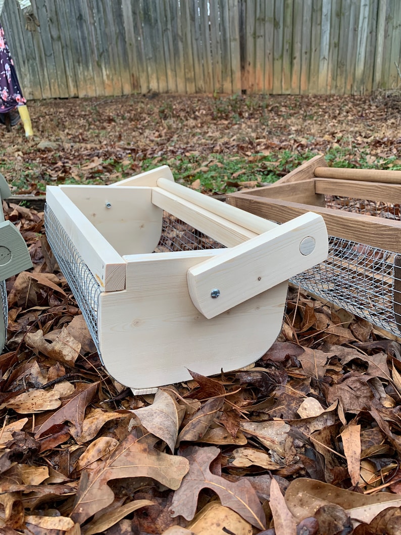 May include: A wooden bird feeder with a wire mesh bottom and a wooden handle. The feeder is sitting on a bed of brown leaves.