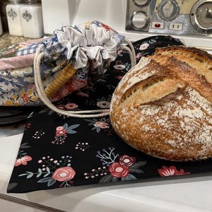 May include: A loaf of freshly baked bread with a scored top rests on a black floral patterned cloth. A drawstring bag and a patchwork fabric bread warmer are also visible, suggesting a kitchen or bakery setting.
