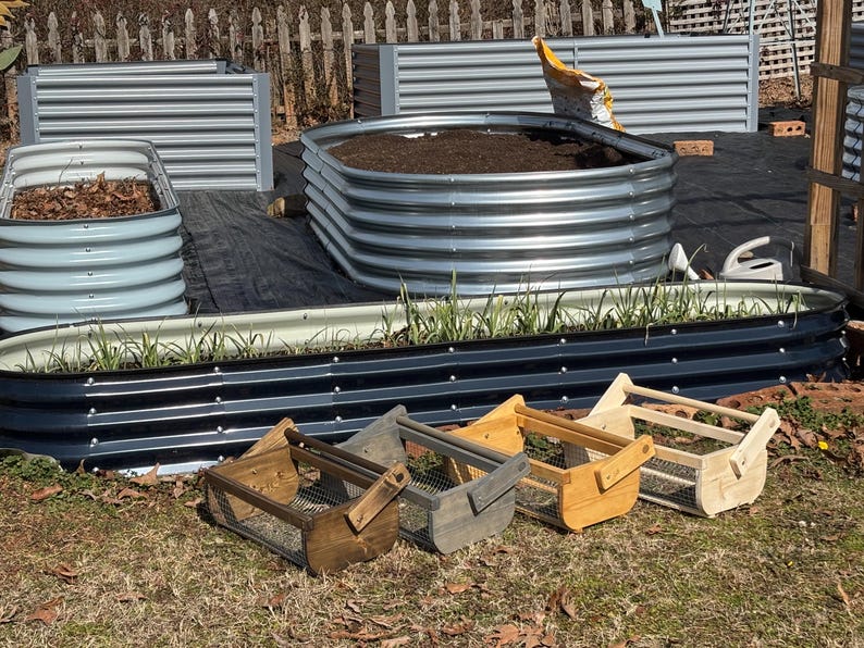 May include: Four wooden baskets with wire mesh bottoms and handles. The baskets are in various shades of brown, gray, and natural wood. The baskets are sitting on a grassy surface in front of a garden.