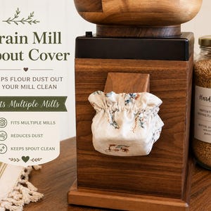 May include: A fabric grain mill spout cover with a floral pattern and elasticized opening, fitted over the wooden spout of a dark wood grain mill. Text on the image reads Grain Mill Spout Cover, Keeps Flour Dust Out & Your Mill Clean, Fits Multiple Mills.