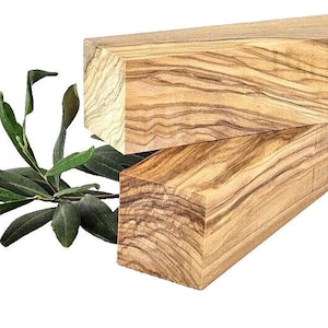 Puede incluir: Dos bloques rectangulares de madera de olivo con patrones de vetas únicos están apilados. Una ramita de hojas de olivo con hojas de color verde oscuro está colocada a la izquierda. La madera es de color marrón claro con vetas marrones más oscuras.
