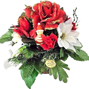 May include: A festive Christmas floral arrangement featuring red roses with gold glitter accents, white poinsettias, and a small Santa figurine. The arrangement includes green leaves and a tag that says "Hand made".