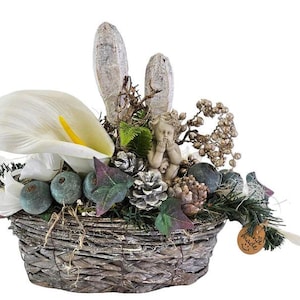 Könnte beinhalten: Dekorativer Korb mit einer weißen Calla-Lilie, grünen Beeren und einer kleinen Cherub-Figur. Der Korb besteht aus geflochtenen Zweigen und enthält Tannenzapfen und dekorative Elemente.