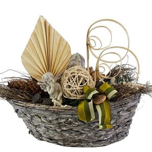 Puede incluir: Un arreglo decorativo en una cesta tejida gris con una pequeña estatua de querubín, piñas y una bola tejida. El arreglo incluye una hoja seca grande en forma de abanico, espirales decorativas y una cinta verde y crema.