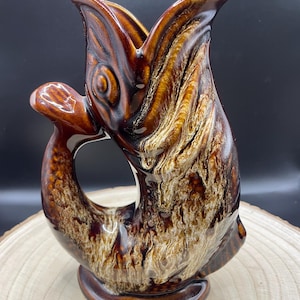 Puede incluir: Jarra de cerámica marrón con forma de pez, con asa y boca abierta. La jarra tiene un acabado brillante con tonos marrones y beige, y se asienta sobre una base redonda. El diseño en forma de pez añade un toque único a esta pieza decorativa.