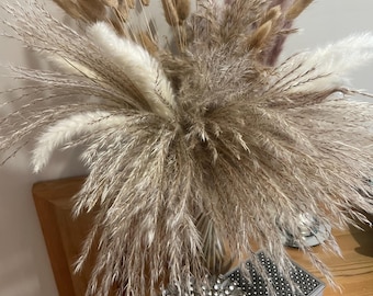 May include: A decorative arrangement of dried pampas grass and other feathery plants in a clear vase. The arrangement includes beige, white, and light brown plumes. A heart-shaped photo frame and a small decorative box sit nearby.