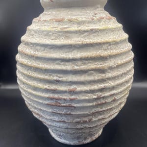 Puede incluir: Un jarrón de cerámica texturizada de color blanquecino, con un cuerpo bulboso y un cuello estrecho. El jarrón presenta detalles horizontales acanalados y zonas de color marrón rojizo expuesto. La estética general es rústica y envejecida.