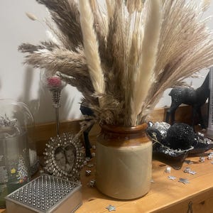 Puede incluir: Un arreglo decorativo de hierba de la Pampa seca y otras plantas plumosas en un jarrón de cerámica marrón y beige. El arreglo está sobre una superficie de madera con otros elementos decorativos, incluyendo una caja plateada y una foto enmarcada.