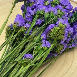 May include: A bundle of dried purple statice flowers with green stems and buds. The flowers are a vibrant purple color, and the stems are long and slender. The flowers are arranged on a light brown wooden surface.