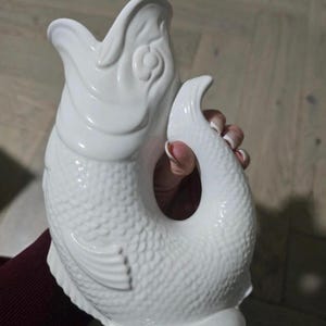 Puede incluir: Jarra blanca de cerámica con forma de pez. La jarra tiene un patrón de escamas texturizadas y un asa curva. La boca del pez está abierta y la cola se curva para formar el asa. La jarra mide aproximadamente 25 cm de alto.