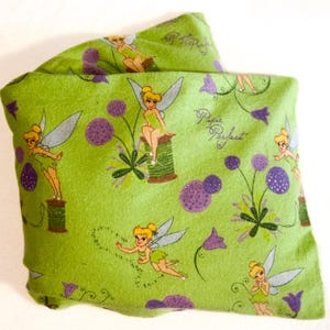 May include: A folded green fabric featuring a repeating pattern of a fairy, purple flowers, and the words "Papie Perfect". The fairy is depicted with blonde hair, a green dress, and silver wings. The fabric is a light green color.