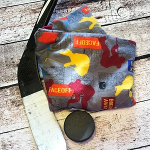 May include: A gray fabric hockey-themed face mask with red, yellow, and black accents. The mask has the words "FACEOFF" and "BREAKAWAY" printed on it. A black hockey puck and a hockey stick are also in the image.