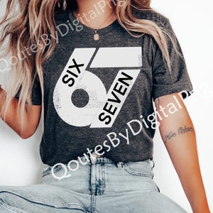May include: Dark gray t-shirt with the text "SIX 67 SEVEN" in a distressed white font. The shirt is a crew neck style and is being worn by a person. The person is wearing blue jeans.