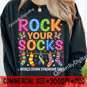 Peut inclure: Sweat-shirt noir avec l'inscription "ROCK YOUR SOCKS" en lettres colorées. Le motif comprend des chaussettes suspendues et un ruban bleu et jaune, avec "WORLD DOWN SYNDROME DAY" en dessous. L'image contient également le texte "COMMERCIAL USE 300DPI PNG".