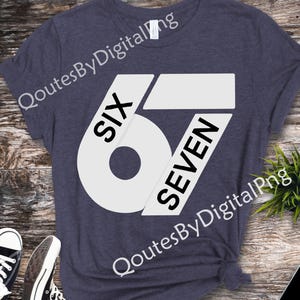 May include: A navy blue t-shirt with a large white graphic design featuring the numbers "67" and the words "SIX" and "SEVEN". The shirt is displayed on a wooden surface with sneakers and a phone.