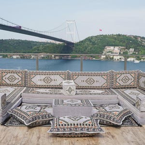 May include: A modular outdoor seating arrangement with a geometric pattern. The set includes cushioned sections, ottomans, and cushions. The colour scheme is grey, beige, and black. The background shows a bridge over water, with a view of the landscape.