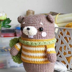 May include: A handmade crochet beaver doll in shades of brown, white, yellow, and green. The beaver has a striped sweater, a green scarf, and pink cheeks. The doll has a white snout with two teeth and black eyes.