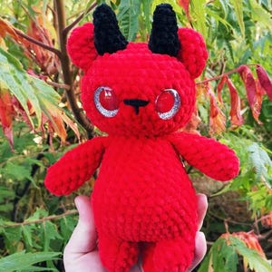 May include: A handmade red plush devil doll with black horns, ears, and hooves. The doll has large, glittery silver eyes and a small black nose. The plushie is held against a backdrop of green and red foliage.
