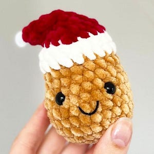 May include: A handmade, crocheted Christmas ornament shaped like a smiling pinecone wearing a Santa hat. The pinecone is tan, with black eyes and a stitched smile. The Santa hat is red with a white trim and a white pom-pom.