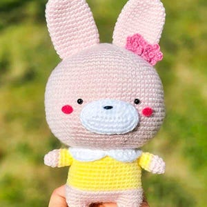 May include: A handmade crochet bunny doll with a pink body, large ears, and a small pink bow. The bunny wears a yellow dress with a white collar and has rosy cheeks. The doll is a soft toy.