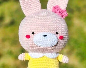 Könnte beinhalten: Handgefertigte Häkelkaninchenpuppe mit rosa Körper, großen Ohren und einer kleinen rosa Schleife. Das Kaninchen trägt ein gelbes Kleid mit weißem Kragen und hat rosige Wangen. Die Puppe ist ein Stofftier.