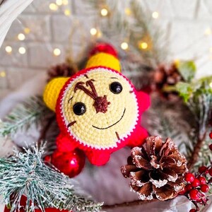 Pode incluir: Um brinquedo de relógio de crochê feito à mão com um rosto sorridente, corpo vermelho, detalhes amarelos e olhos pretos. O relógio é cercado por decorações festivas, incluindo pinhas, enfeites vermelhos e galhos de pinheiro artificiais.