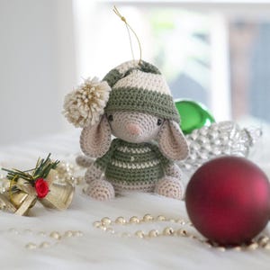 Peut inclure: Ornement de Noël fait à la main en crochet en forme de souris. La souris porte un chapeau et un pull rayés verts et blancs, avec un pompon et des oreilles beiges. Une boule de Noël rouge et des cloches dorées sont en arrière-plan.