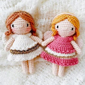 May include: Two handmade crochet angel dolls. One doll wears a white dress with gold trim, the other a pink dress with white and gold trim. Both dolls have yarn hair, rosy cheeks, and small wings.