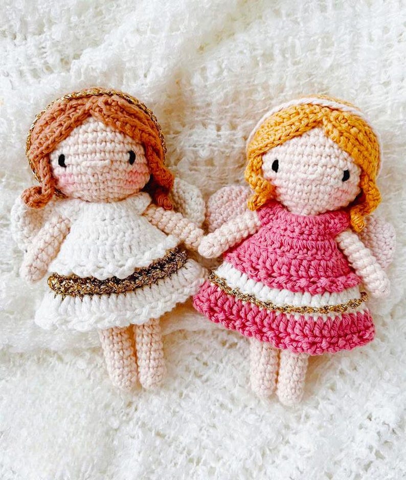 May include: Two handmade crochet angel dolls. One doll wears a white dress with gold trim, the other a pink dress with white and gold trim. Both dolls have yarn hair, rosy cheeks, and small wings.