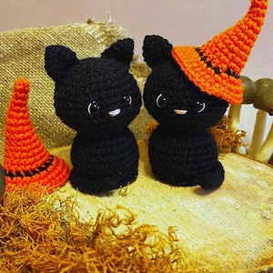 Può includere: Figurine di gatti neri all'uncinetto con occhi e baffi bianchi. Un gatto indossa un cappello da strega arancione con una fascia nera. L'altro gatto non ha il cappello. Le figurine sono su una superficie di legno con muschio arancione, perfette per Halloween.