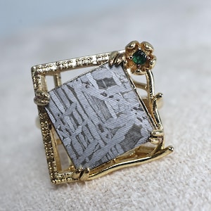 May include: A gold-toned ring featuring a square, patterned, gray stone set within a square frame. A small gold flower with a green gemstone accent is on the side. The ring has a unique, artistic design.