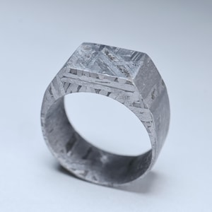 May include: A silver-colored signet ring with a geometric design. The ring has a square top and a thick band. The metal has a unique pattern, with visible lines and textures. The ring is likely made of a durable material.