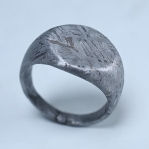 Puede incluir: Primer plano de un anillo de sello plateado. El anillo tiene una superficie texturizada con un patrón en la parte superior. El anillo es redondo con una parte superior plana y una banda que rodea el dedo.