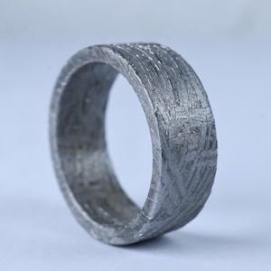 Puede incluir: Un anillo de metal gris que presenta un patrón de Widmanstätten distintivo en toda su superficie. La banda tiene un perfil ancho y plano con un acabado texturizado y grabado que muestra líneas cristalinas que se cruzan por el exterior e interior.