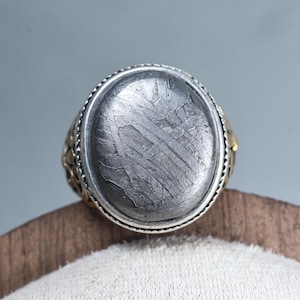Puede incluir: Anillo ovalado con una banda plateada y una piedra gris texturizada. El anillo tiene un acento decorativo de color dorado en el lateral. La piedra tiene un patrón estriado único.