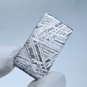 May include: A rectangular slice of iron meteorite featuring a distinct Widmanstätten pattern of intersecting metallic bands. The surface displays a polished, silvery-gray finish with a complex, geometric crystalline structure across the entire face.