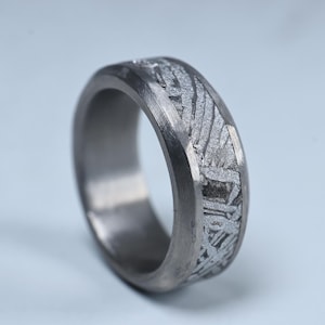 May include: A silver ring with a textured pattern. The ring has a wide band and a matte finish. The pattern is a unique, geometric design. The ring is likely made of metal. The ring's diameter is approximately 2 cm.
