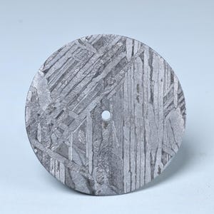 Op de afbeelding: Een cirkelvormig, metalen object met een complex, getextureerd patroon. Het oppervlak toont een netwerk van elkaar kruisende lijnen en vormen, met een klein gaatje in de buurt van het midden. Het object heeft een zilver-grijze kleur en lijkt van metaal te zijn.