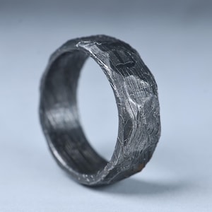 Può includere: Un anello grigio scuro con una superficie ruvida e fortemente strutturata che ricorda la corteccia o la pietra. La fascia presenta un bordo esterno organico e irregolare con solchi e creste profonde su tutto il metallo, creando un aspetto opaco ossidato.