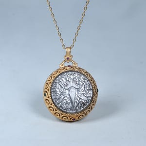 Könnte beinhalten: Eine goldfarbene Anhängerkette mit detailliertem kreisförmigem Design. Der Anhänger hat eine silberfarbene Mitte mit einem baumartigen Design, umgeben von einem goldenen Filigranrand. Kleine rote und grüne Edelsteine sind am Rand eingesetzt. Die Halskette ist ebenfalls goldfarben.