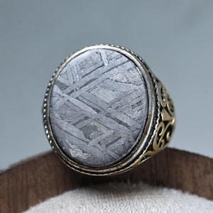 Peut inclure: Bague ovale avec une pierre grise à motifs, sertie dans une lunette argentée. La bague a une bande décorative dorée avec un design orné. Le motif de la pierre a un aspect géométrique unique.