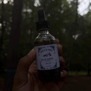 May include: A clear glass bottle with a black dropper, filled with a dark liquid and green herbs. The label reads "Seriya & Smoke" and "Black Dahlia Spell Oil". The bottle is held against a blurred green background.