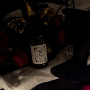 May include: A dimly lit image showing a bottle of "Catwoman" spell oil. The label is white with black text and floral designs. The bottle is set amongst dark fabrics, gold decorations, and red rose petals.