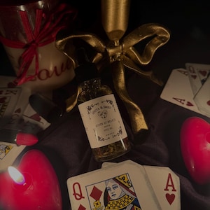 May include: A bottle of "Queen of Hearts" spell oil with a dropper, surrounded by playing cards, a candle, and a gold-coloured decorative bow. The label features floral accents and the brand name "Seriya & Smoke."